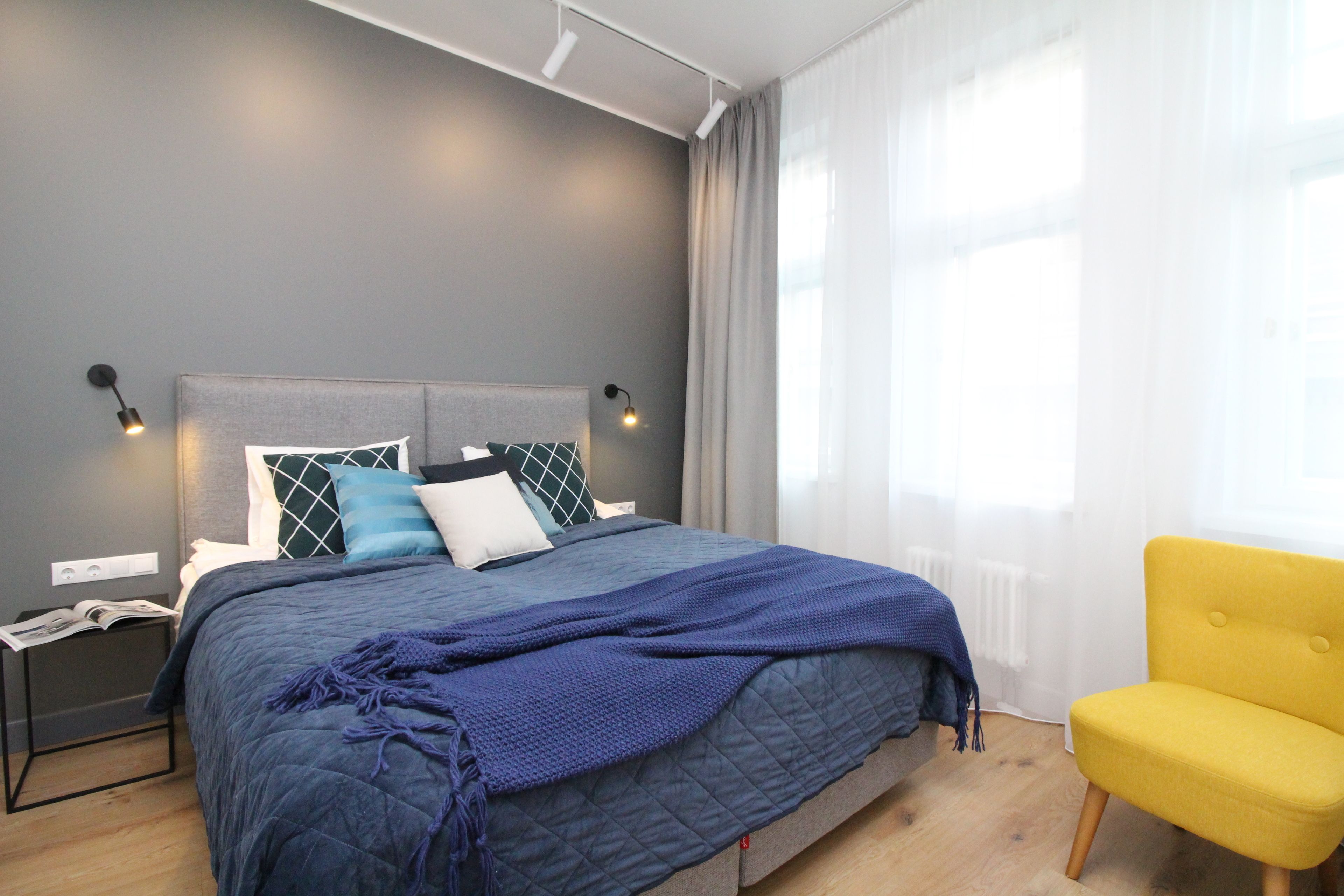 A modern bedroom with a large bed, a nightstand, a yellow armchair, and a window with sheer curtains.