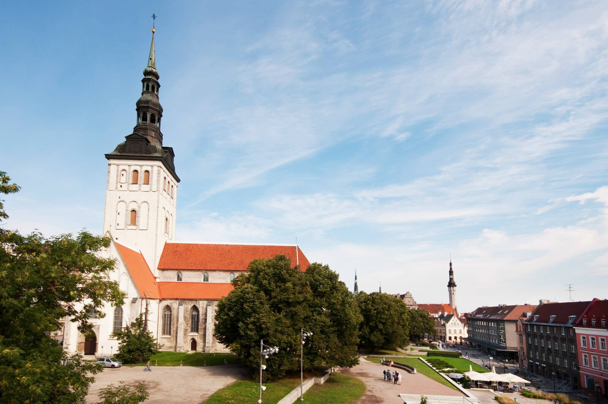 A historic church with a prominent tower and a red roof, surrounded by trees and a paved square.