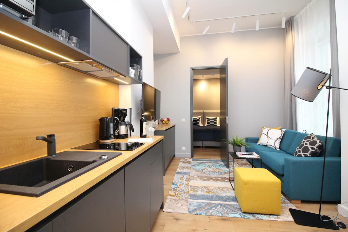Cozinha moderna com pia preta, bancada de madeira e cafeteira. Uma sala de estar com sofá azul e um banquinho amarelo também é visível.