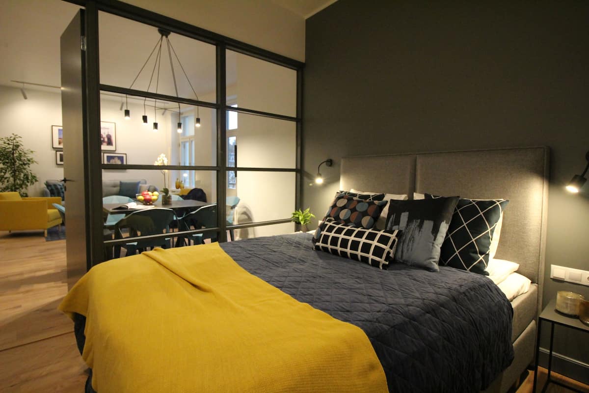 Quarto moderno com uma cama grande, um cobertor amarelo e vista para uma sala de jantar através de uma divisória de vidro.
