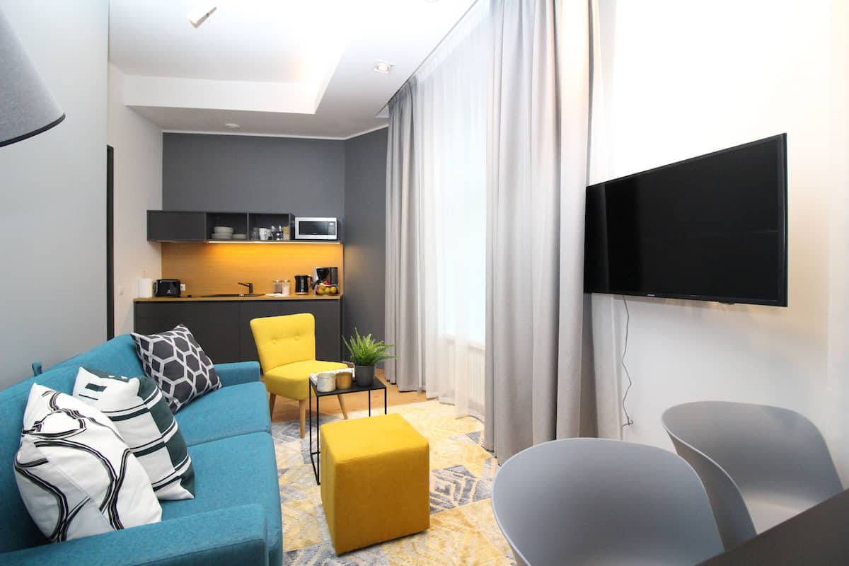 Uma sala de estar moderna com sofá, banquinho e poltronas, uma mesinha com uma planta, e uma área de cozinha com micro-ondas.