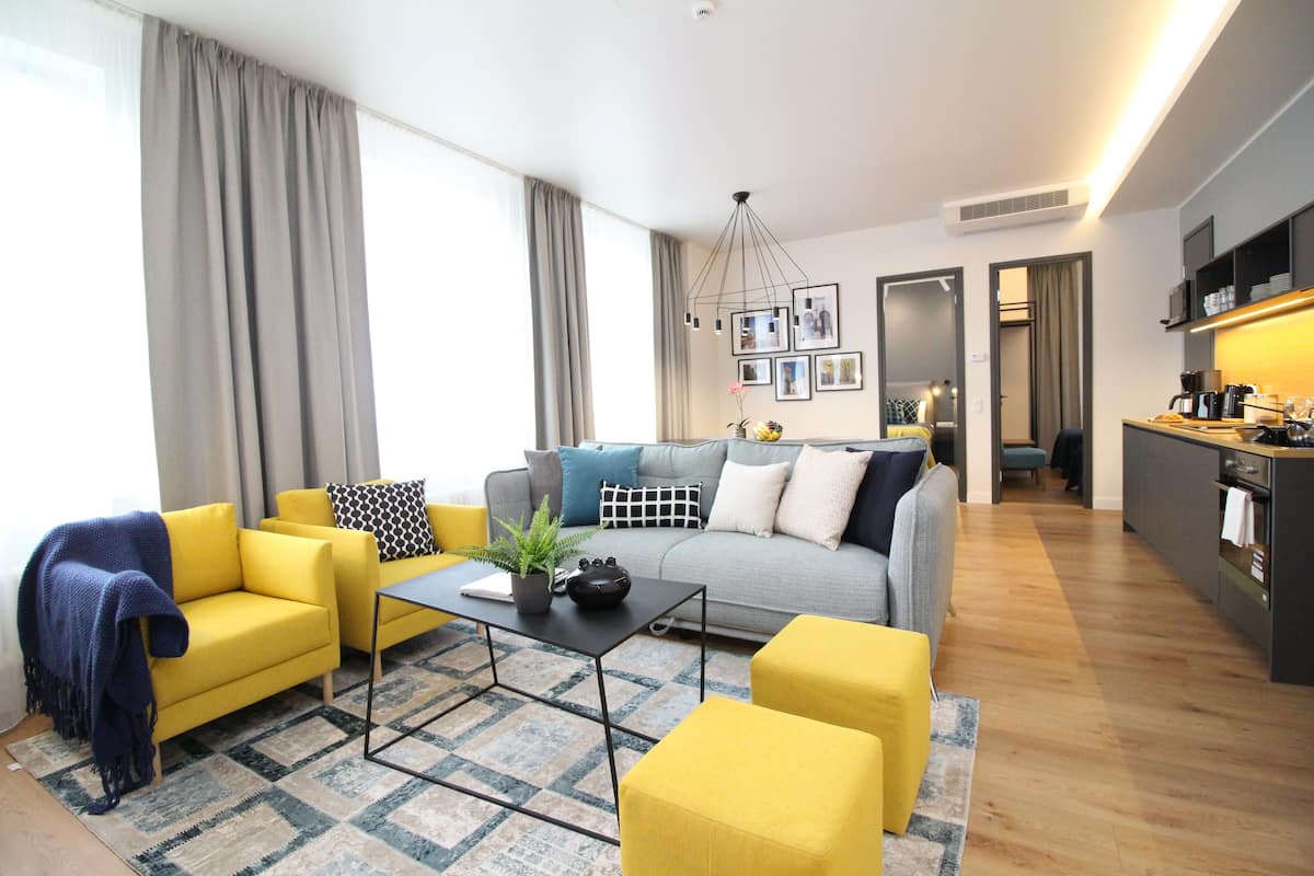 Uma sala de estar moderna com um sofá, poltronas e uma mesa de centro. Há uma área de cozinha com bancada e eletrodomésticos.