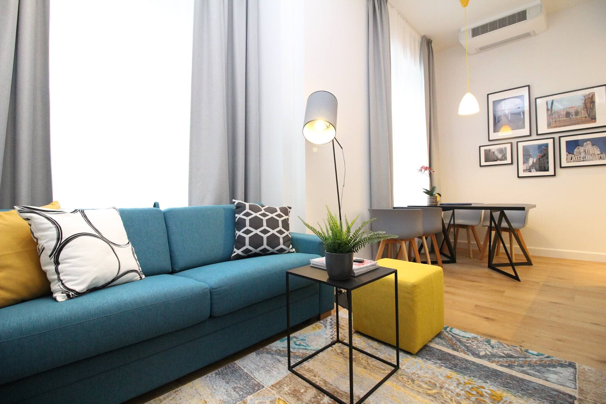 Un salón con un sofá azul, una pequeña mesa negra con una planta, un reposapiés amarillo y un comedor con una mesa y sillas.