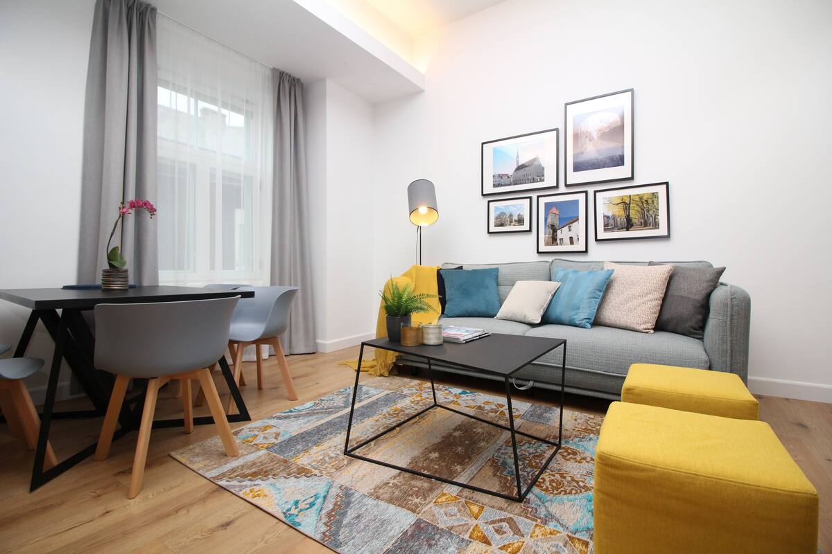 Sala de estar moderna com sofá cinza, um banquinho amarelo e uma mesa de centro. Há uma mesa de jantar com cadeiras e um abajur. Quadros pendem na parede.