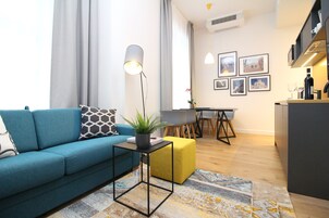 Uma sala de estar moderna com um sofá azul, uma mesinha lateral com uma planta e um banquinho amarelo. Ao fundo, há uma área de jantar com uma mesa e cadeiras.