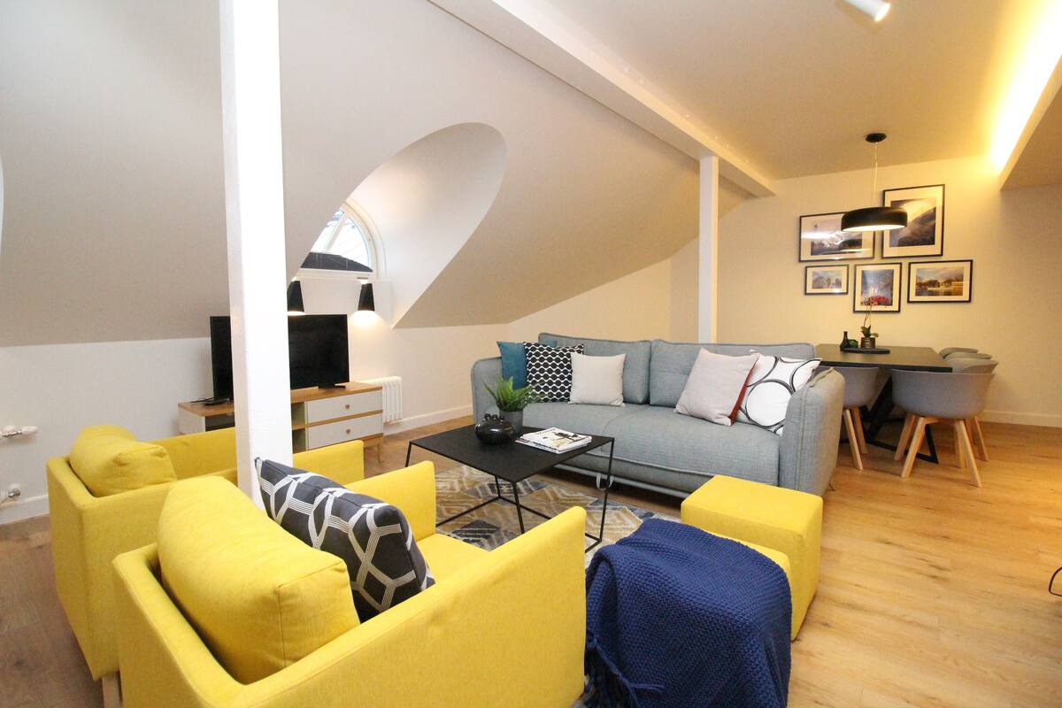 Uma sala de estar moderna com um sofá cinzento, poltronas amarelas e uma mesa de centro. Há também uma área de jantar com uma mesa pequena e cadeiras, e uma TV sobre um suporte.