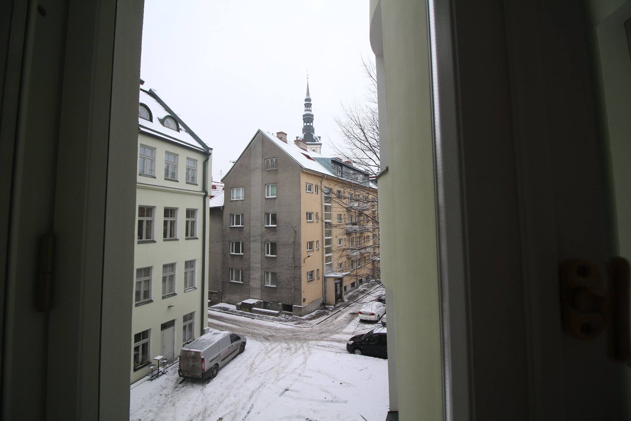 Een uitzicht door een open deur op een besneeuwde stadstraat, met gebouwen en een kerktoren op de achtergrond.