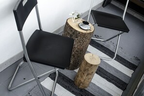 Ein schwarzer Stuhl mit Metallgestell, ein kleiner runder Holztisch mit einer Glasvase und einer Tasse, und ein Streifen Teppich auf Fliesenboden.