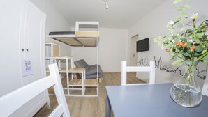 Een kleine kamer met een stapelbed, een klein tafeltje met een stoel, een aan de muur bevestigde televisie en een vaas met bloemen op een tafel.