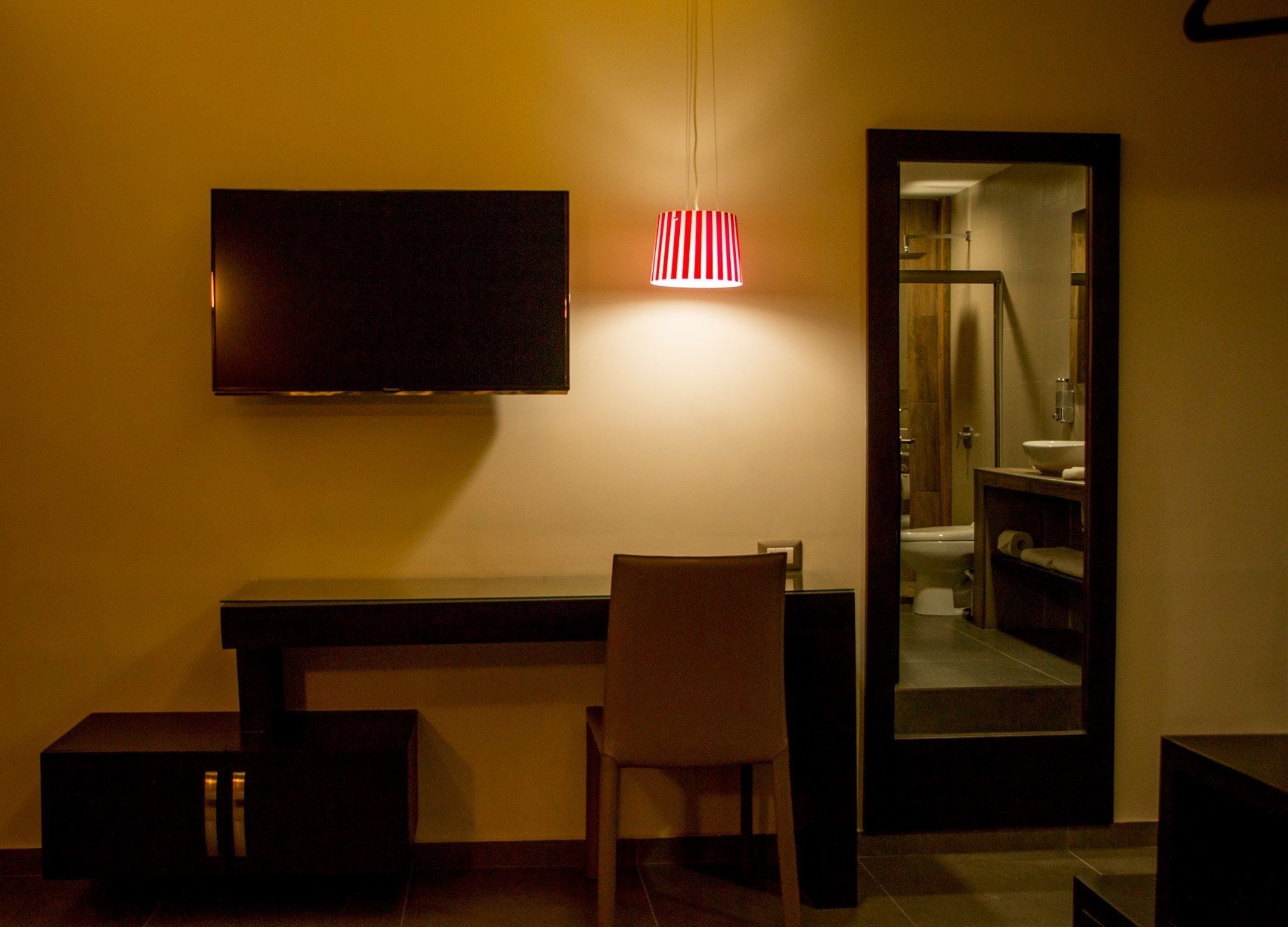 Habitación de hotel con televisión de pantalla plana en la pared, un pequeño escritorio, una silla y un espejo que refleja un baño.