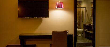 A hotel room with a flat-screen TV mounted on the wall, a small desk, a chair, and a mirror reflecting a bathroom.