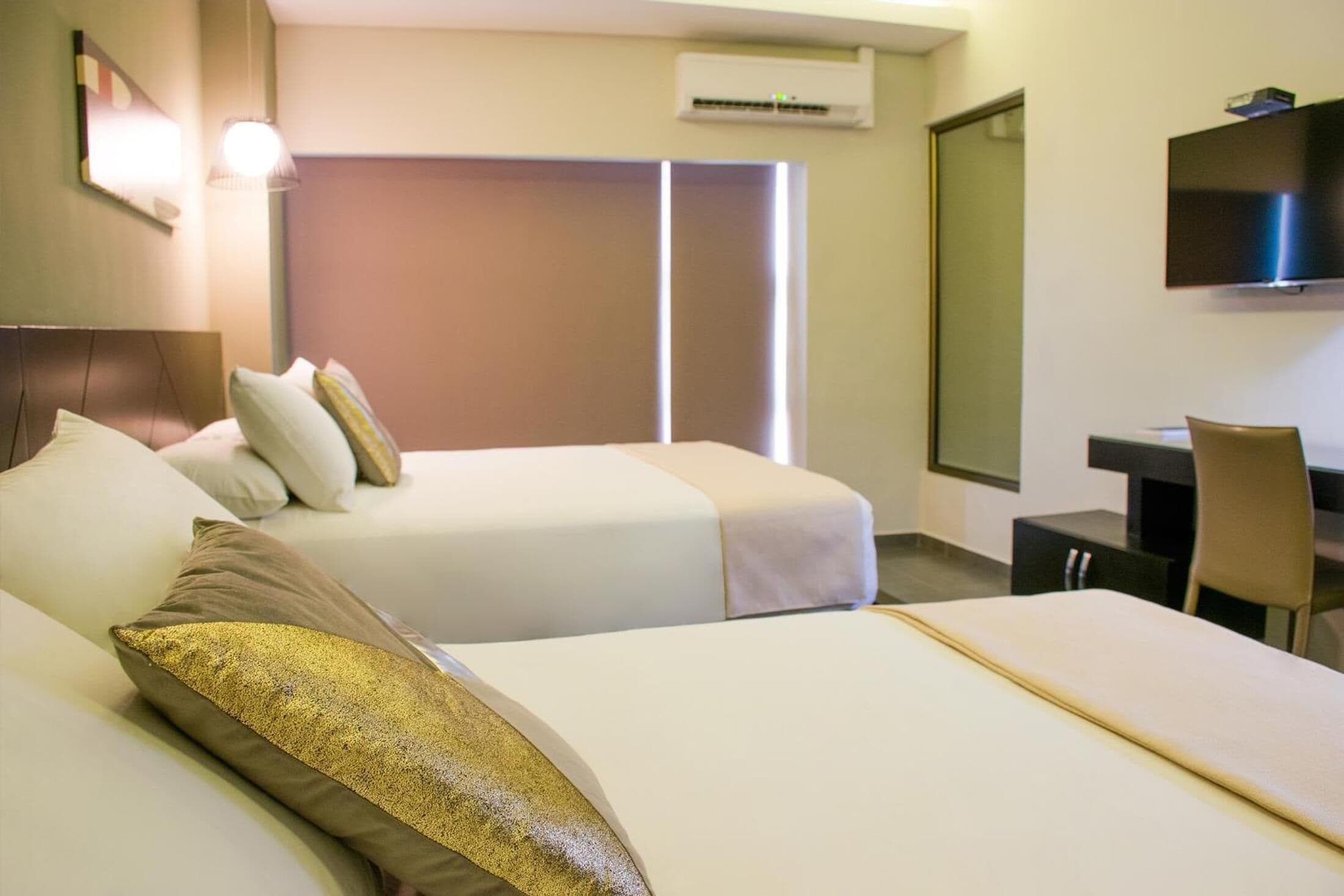 A hotel room with two beds, a desk, a chair, a TV, and an air conditioning unit.