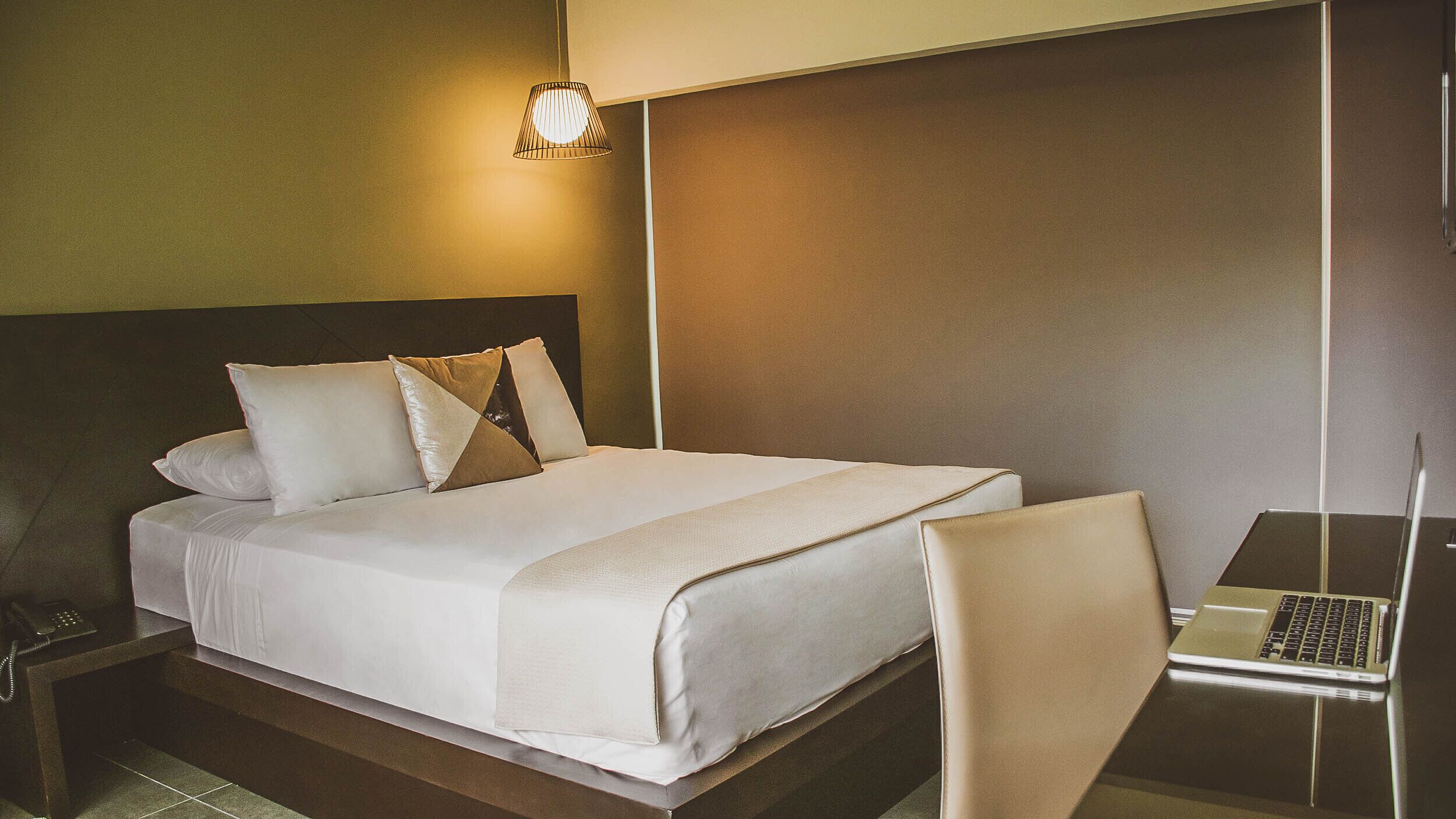 A hotel room with a bed, a desk with a laptop, a telephone, and a wall-mounted light.