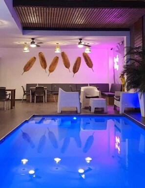 A modern indoor pool area with a blue-lit pool, ceiling fans, and wicker lounge chairs. - Hotel Remfort (Santa Ana)