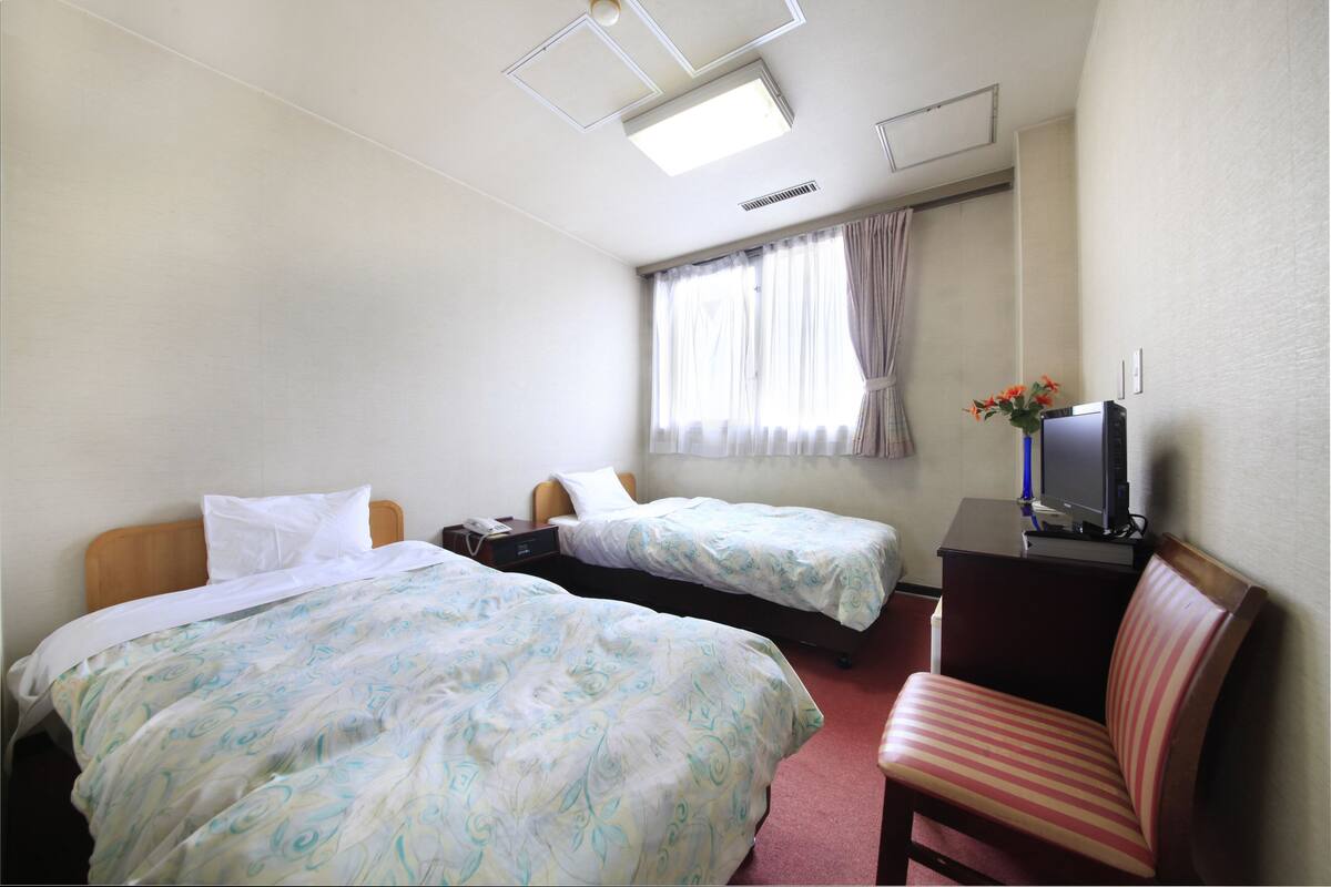A hotel room with two beds, a desk, a chair, a TV, and a window with curtains.