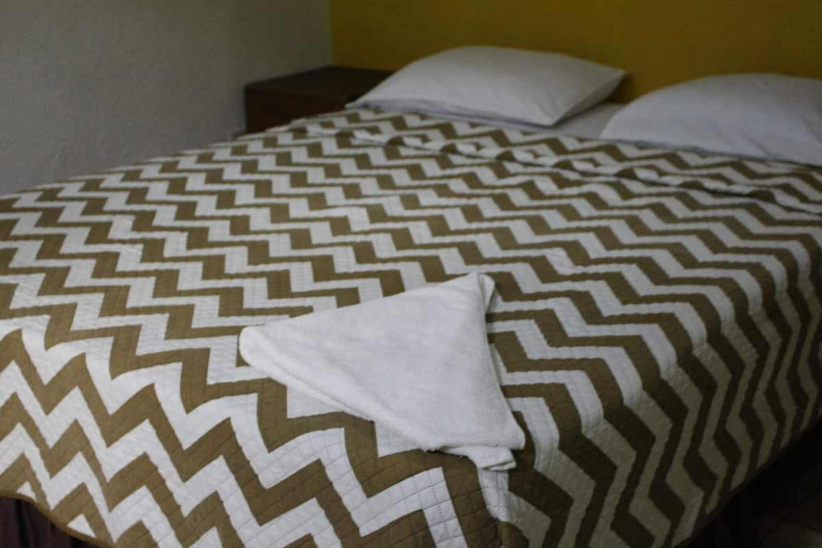 Una cama con una colcha estampada y almohadas, una toalla blanca sobre la cama y una mesita de noche con una lámpara.