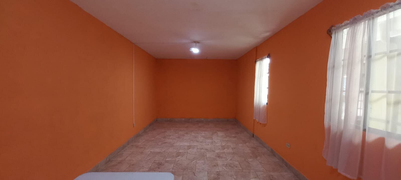 Una habitación con paredes anaranjadas, piso de baldosas, una sola ventana con cortina blanca y una lámpara en el techo.