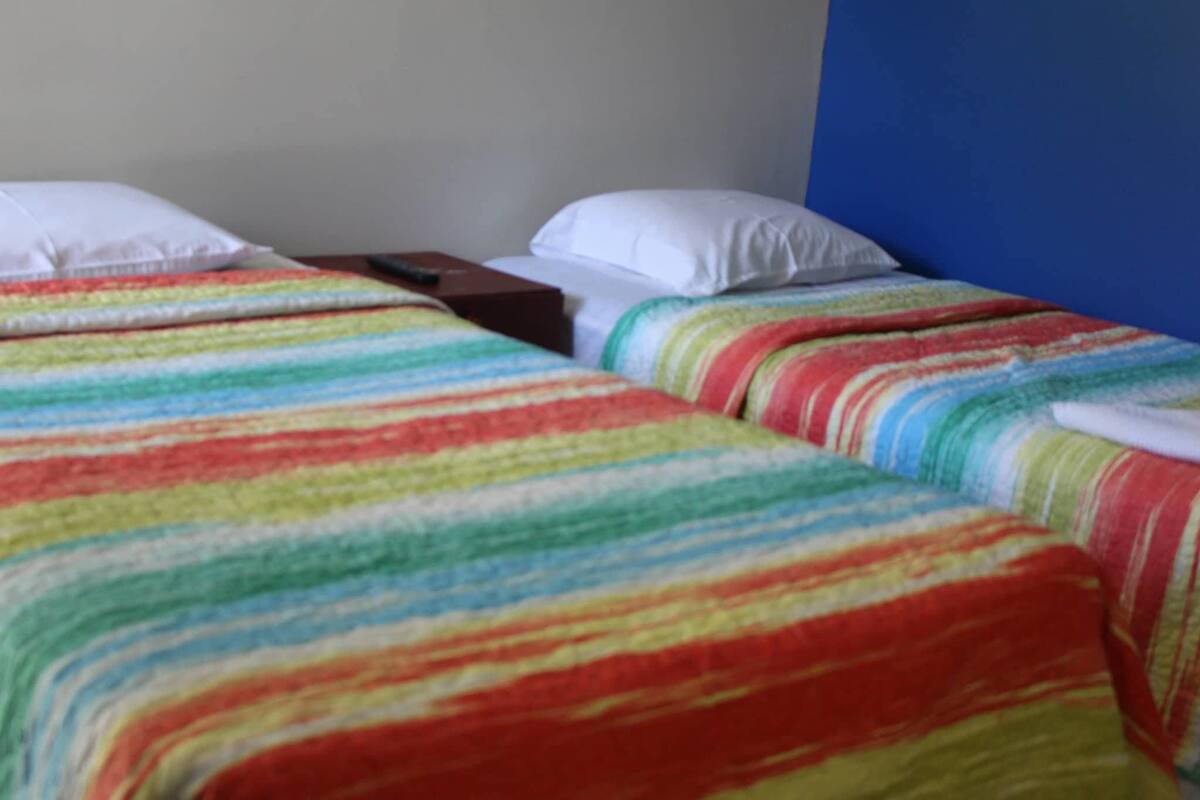 Habitación de hotel con dos camas individuales, cada una con una manta a rayas de colores y almohadas blancas.