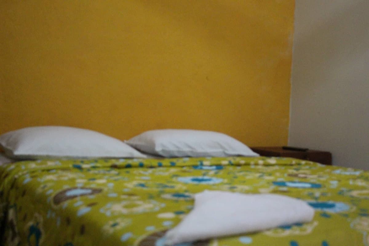 Una habitación con una cama cubierta por una colcha estampada con motivos florales, un almohadón blanco y una pared amarilla.