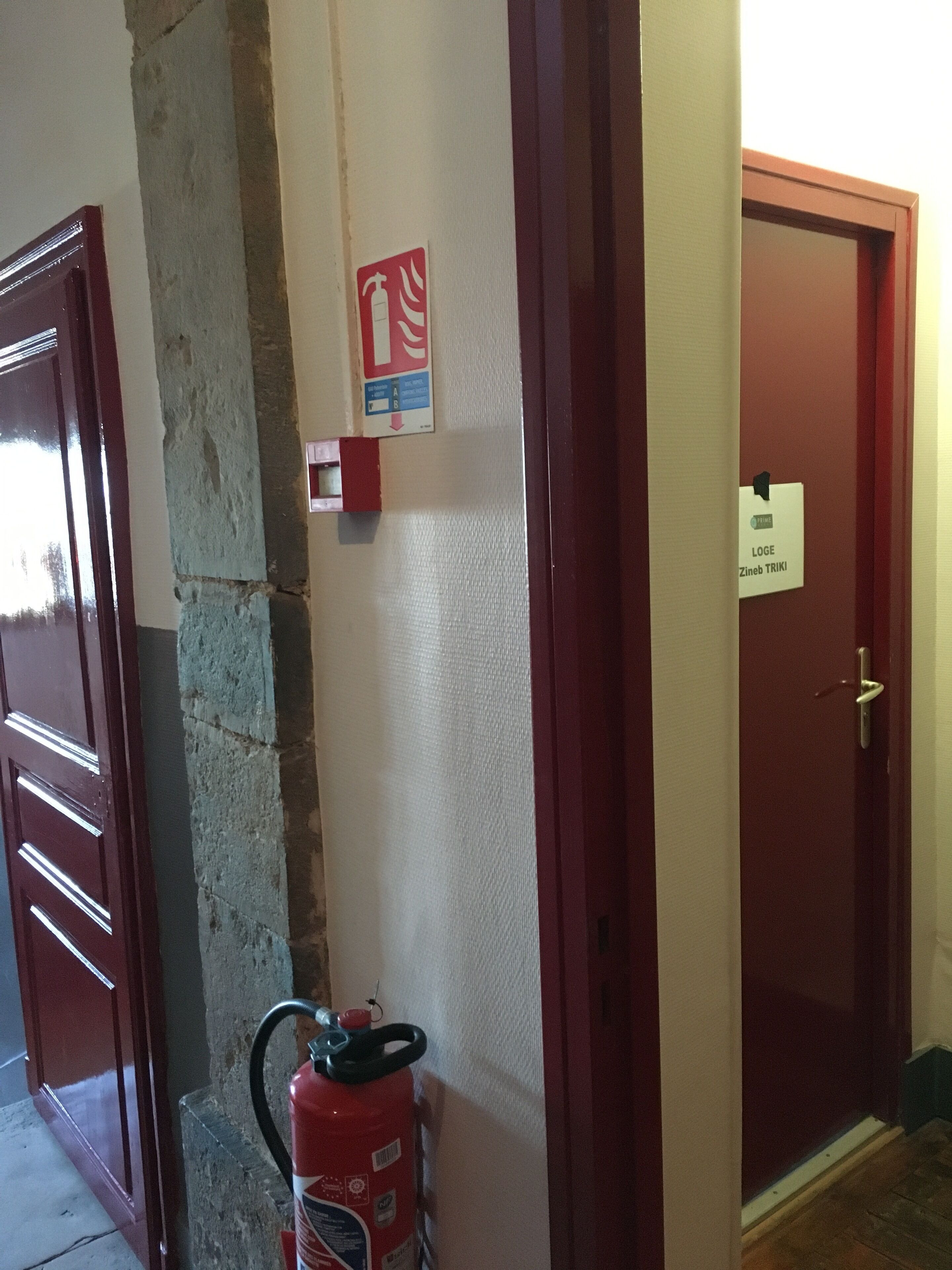 A hallway with a fire extinguisher, a fire safety sign, and a door labeled "LOGE Zineb TRIKI".
