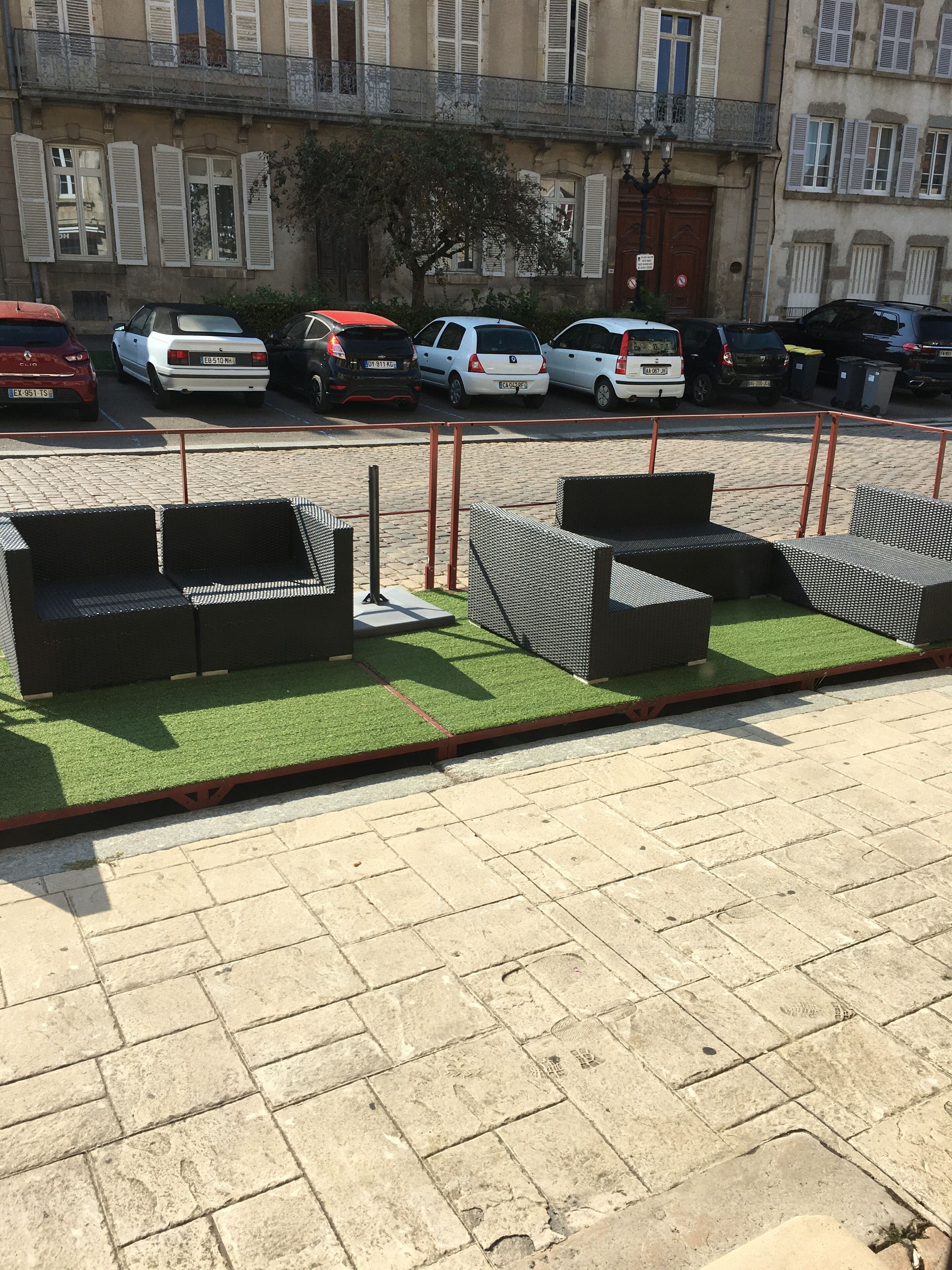 Mobiliario exterior con césped artificial y estructura metálica, un camino pavimentado y coches estacionados al fondo.