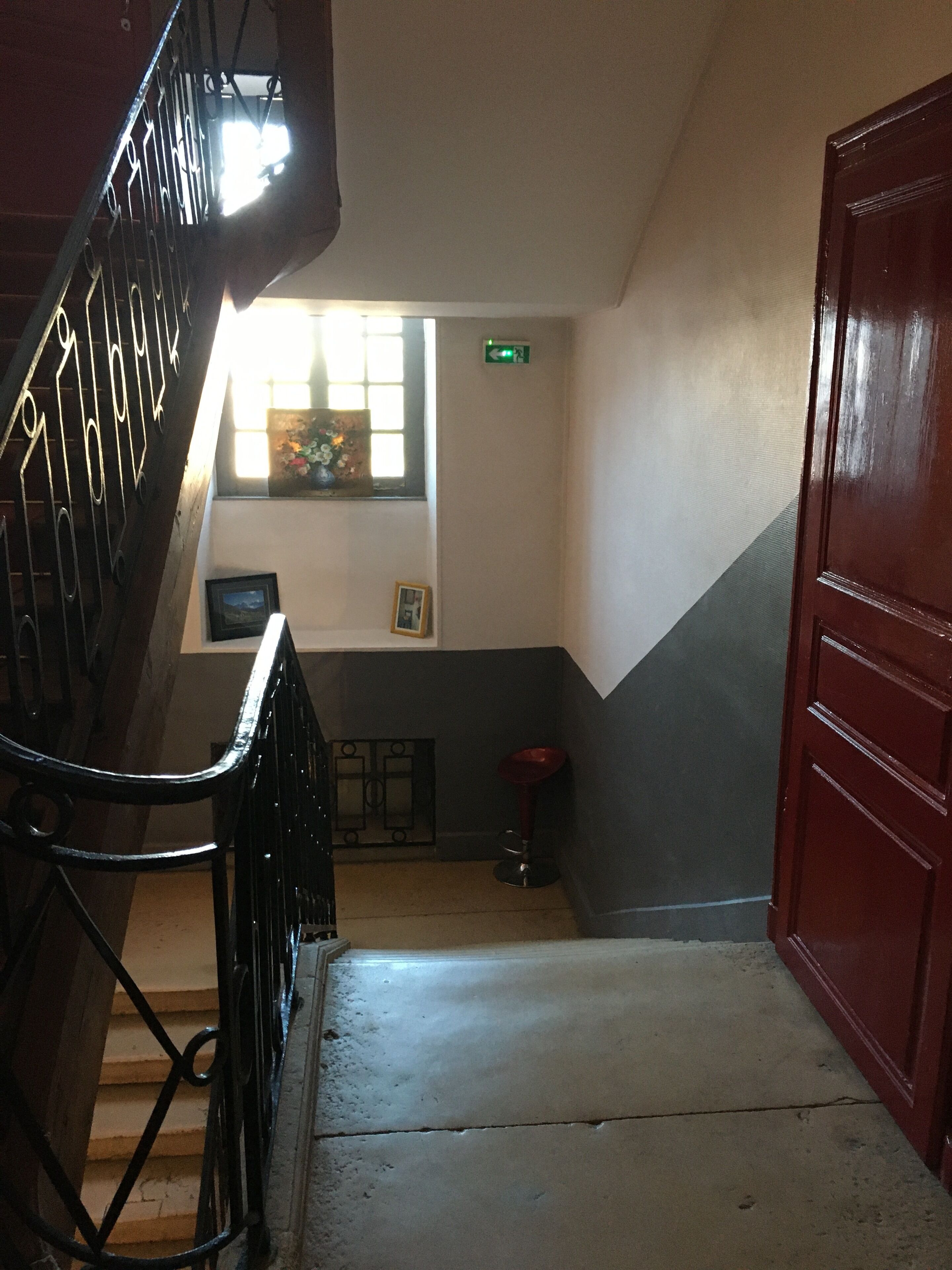 Una escalera con barandilla que conduce a una plataforma. Se observa una ventana con vistas al exterior, una puerta en la plataforma y un taburete pequeño.