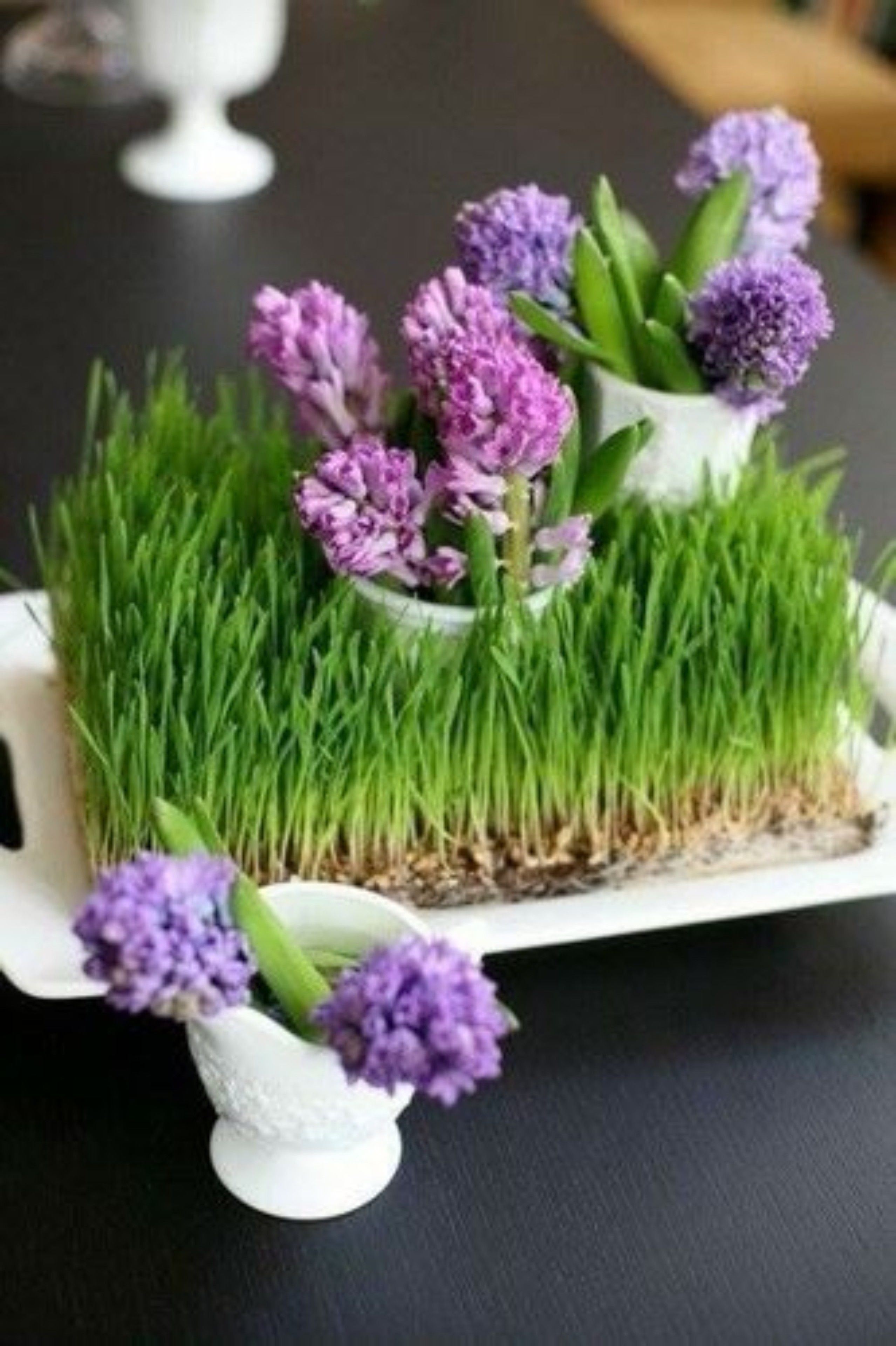 A white ceramic pot with purple flowers and green grass on a black surface.
