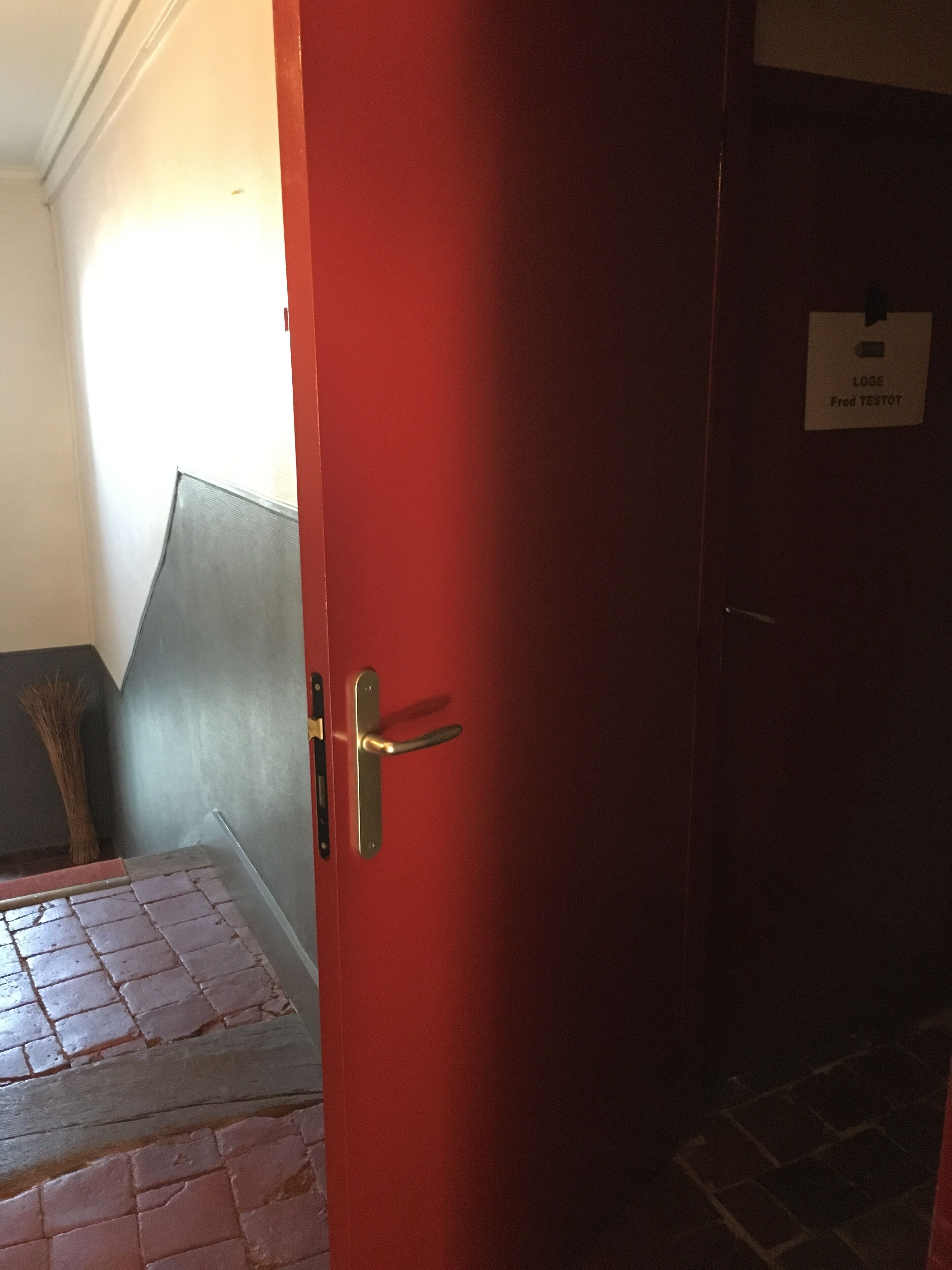 A red door with a brass handle, a sign reading "LOGE Fred TESTOT", and a broom leaning against the wall.