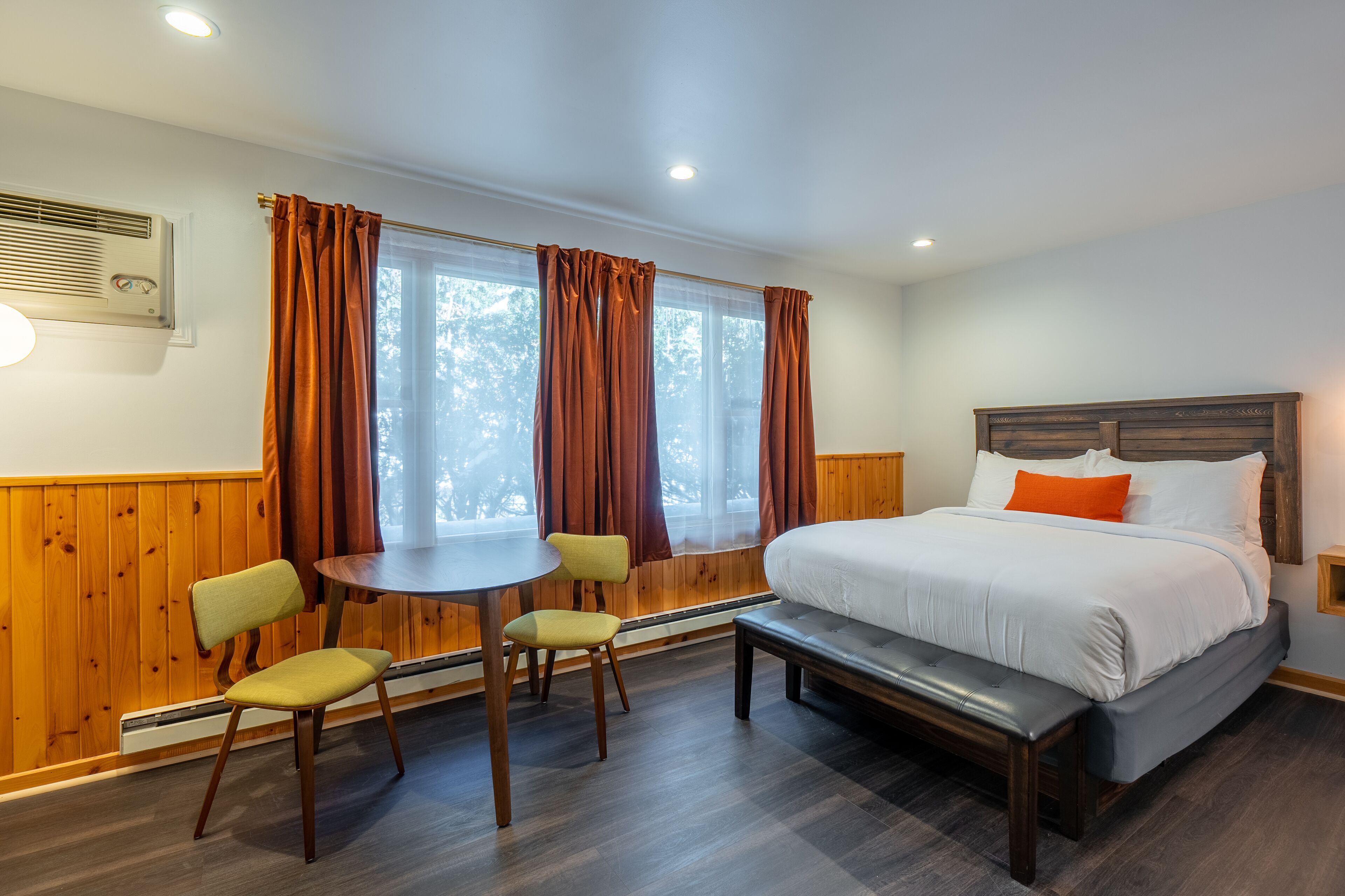 A bedroom with a bed, a bench, a table, two chairs, and a window with curtains.