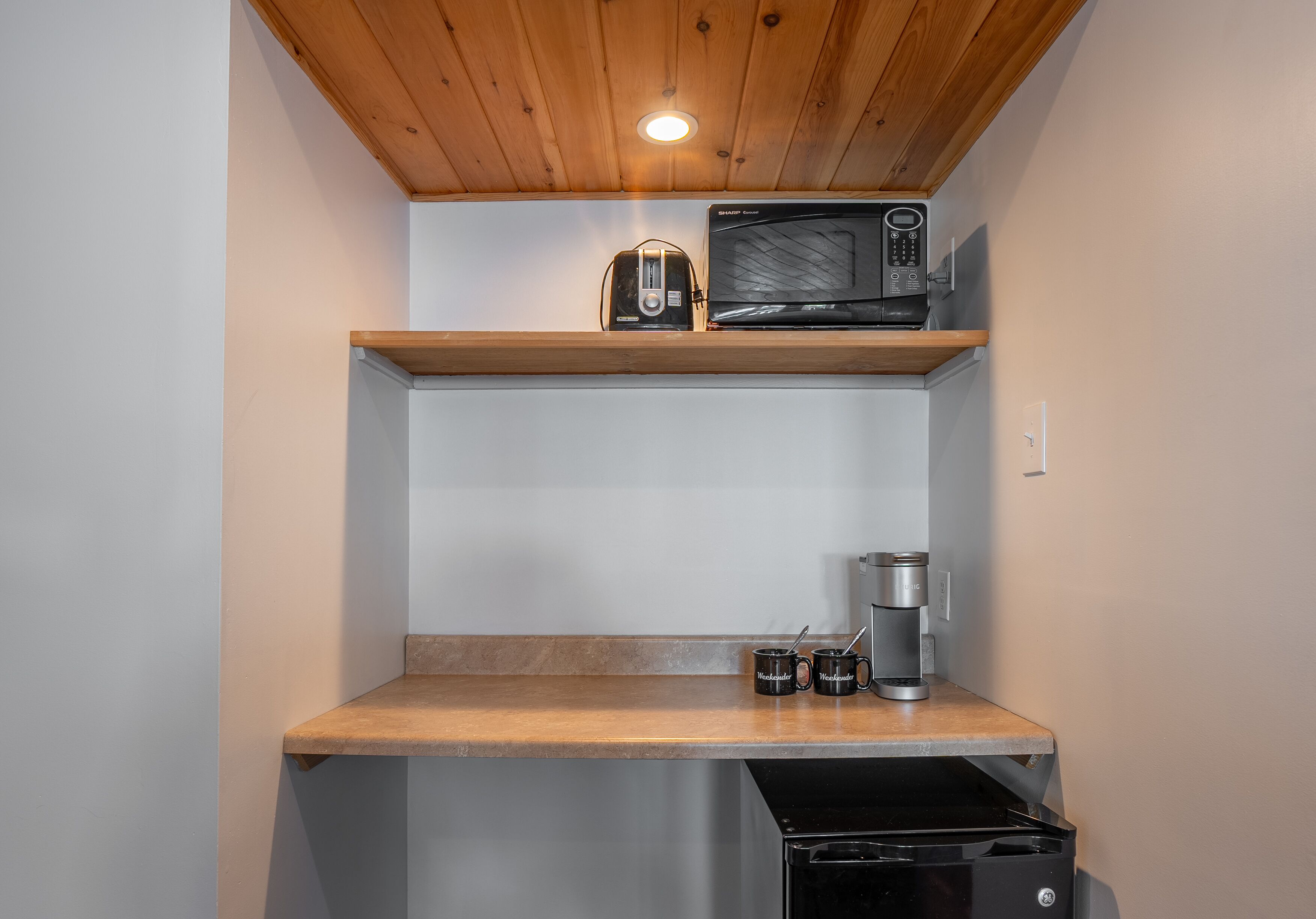 Una kitchenette compacta con microondas, tostadora y cafetera sobre un estante de madera, y un cubo de basura debajo.