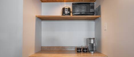 Una kitchenette compacta con microondas, tostadora y cafetera sobre un estante de madera, y un cubo de basura debajo.