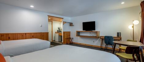 A hotel room with two beds, a desk, a chair, a television, and a small kitchenette.