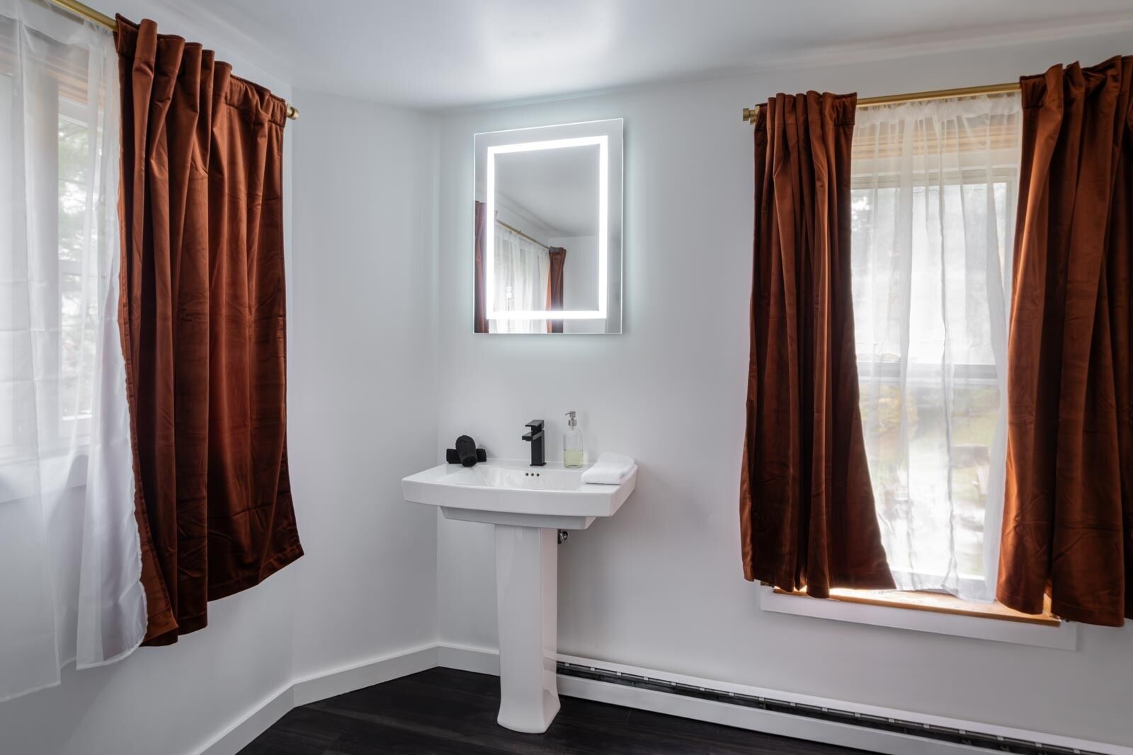 A bathroom with a white pedestal sink, a mirror above it, and brown curtains on the windows.