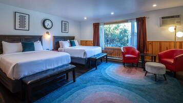 A hotel room with two beds, a bench, a red armchair, a small ottoman, a clock, and a window with curtains.