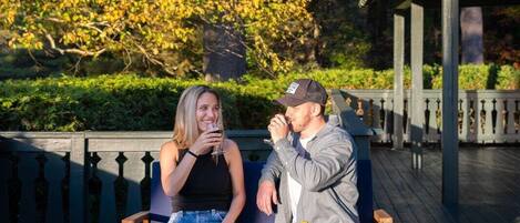 Two people sitting on a wooden bench outdoors with a bottle of wine on the table.