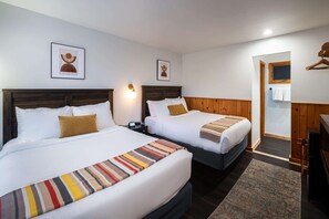 A hotel room with two beds, a wooden headboard, a small desk, a nightstand, a wall-mounted light, and a door leading to a bathroom. - The Town House Lodge (Lake Placid)