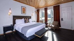 Une chambre moderne avec un grand lit, des tables de chevet, un banc, une chaise et une vue sur l’extérieur grâce à de grandes fenêtres.