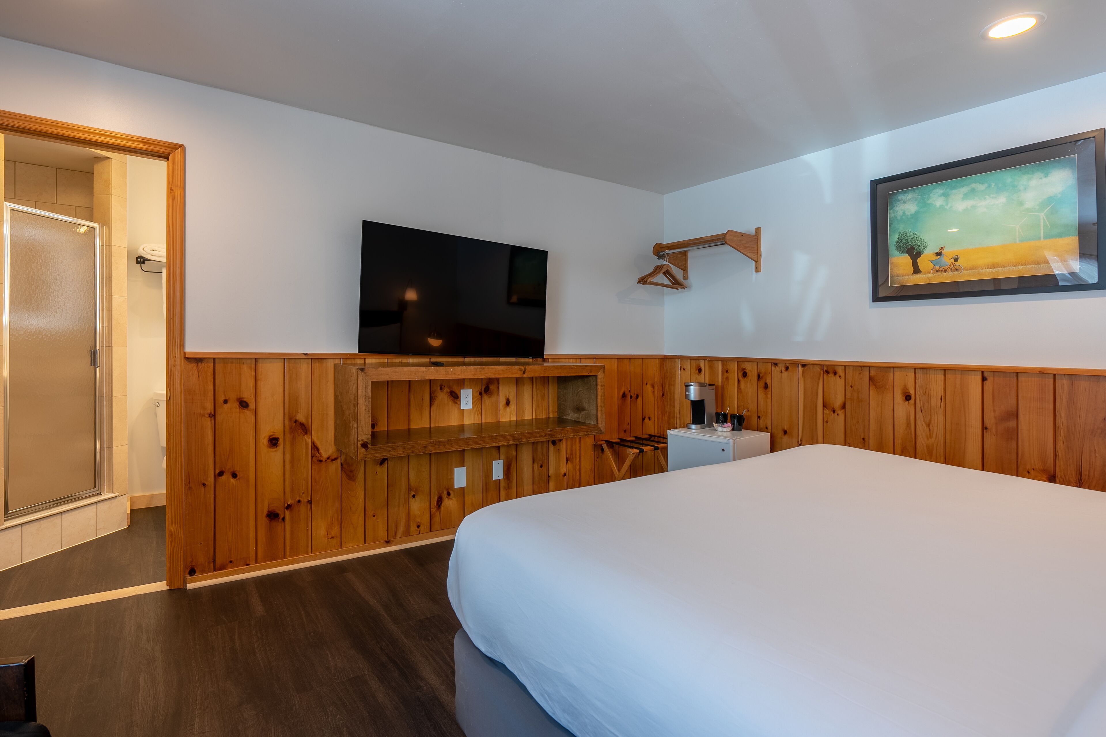 Un dormitorio con una cama grande, televisión montada, paneles de madera, un cuadro enmarcado y una mesita de noche pequeña.