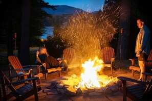 A campfire with chairs around it, a person standing, and another person sitting. - The Town House Lodge (Lake Placid)