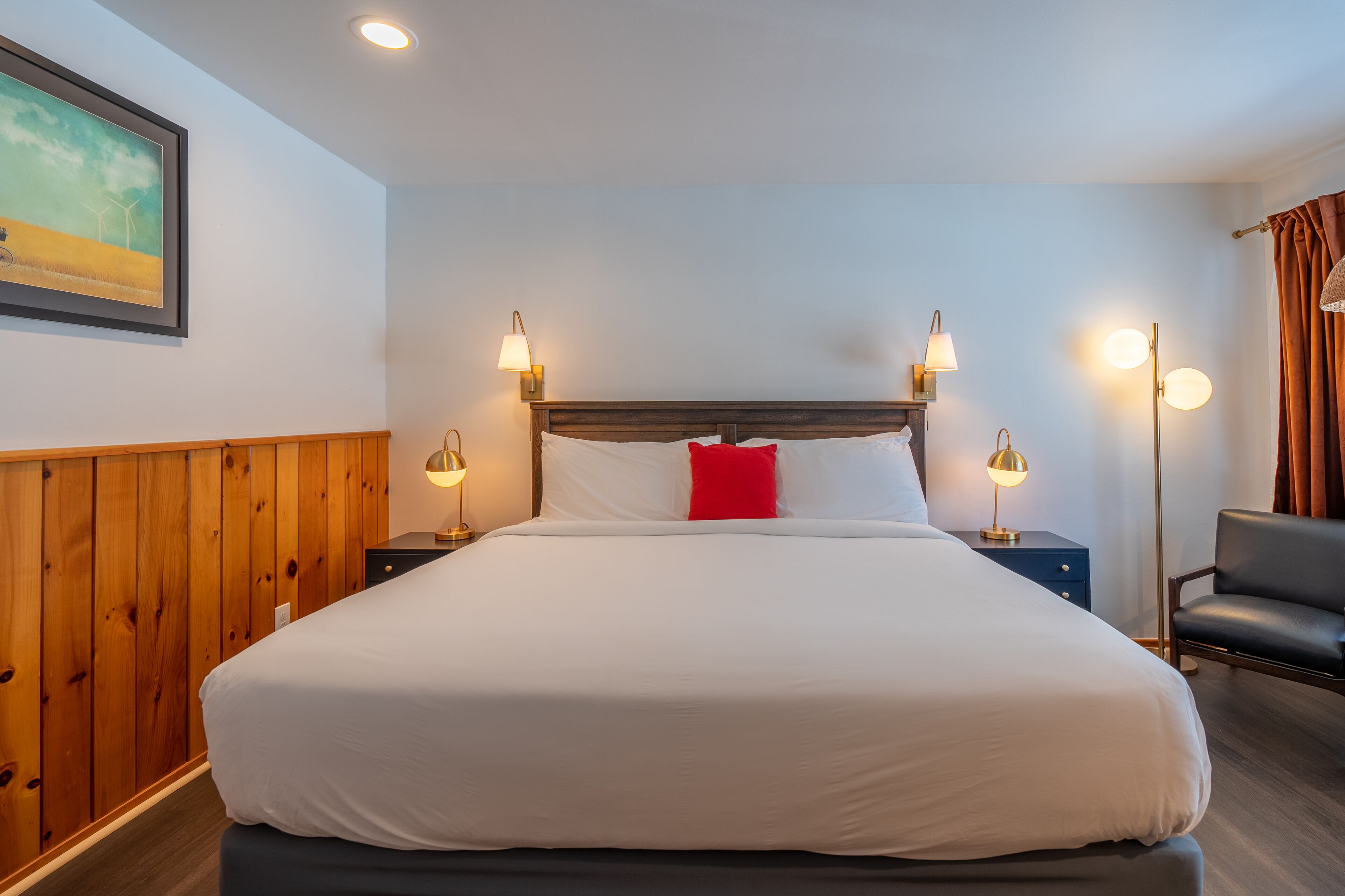 Ein großes Bett mit weißer Bettwäsche, zwei Nachttischlampen, ein rotes Kopfkissen und ein hölzernes Kopfteil.