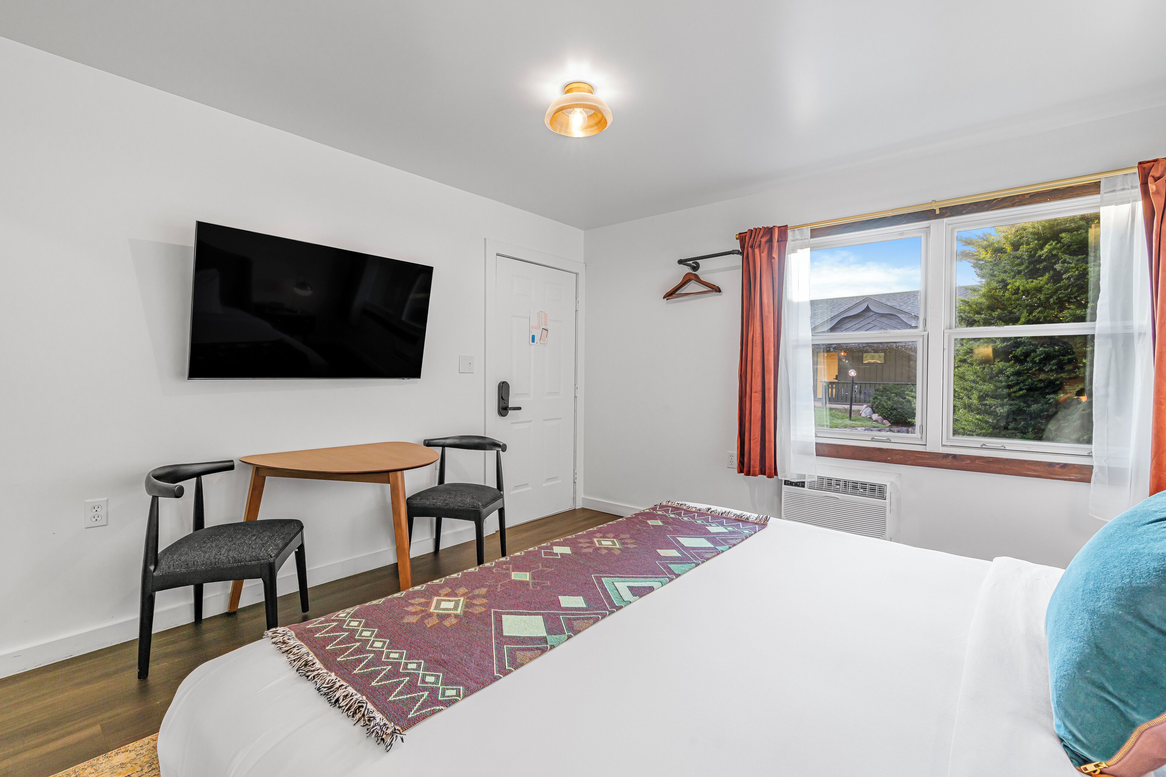 A hotel room with a bed, a TV mounted on the wall, a small table with two chairs, a window with curtains, and a patterned rug.