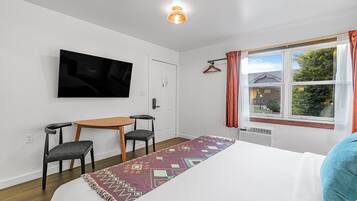 A hotel room with a bed, a TV mounted on the wall, a small table with two chairs, a window with curtains, and a patterned rug.
