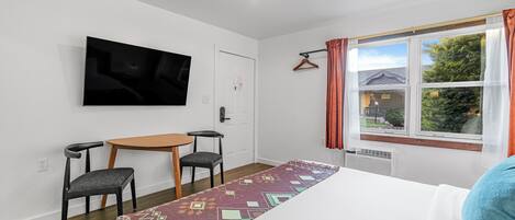 A hotel room with a bed, a TV mounted on the wall, a small table with two chairs, a window with curtains, and a patterned rug.