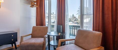Una habitación con dos sillones de cuero marrón, una mesita redonda con mantel negro, una mesita auxiliar negra con cajón y una ventana con cortinas rojas.