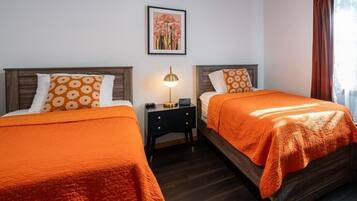 A hotel room with two beds, each with an orange bedspread and matching pillows. There is a nightstand with a lamp between the beds.
