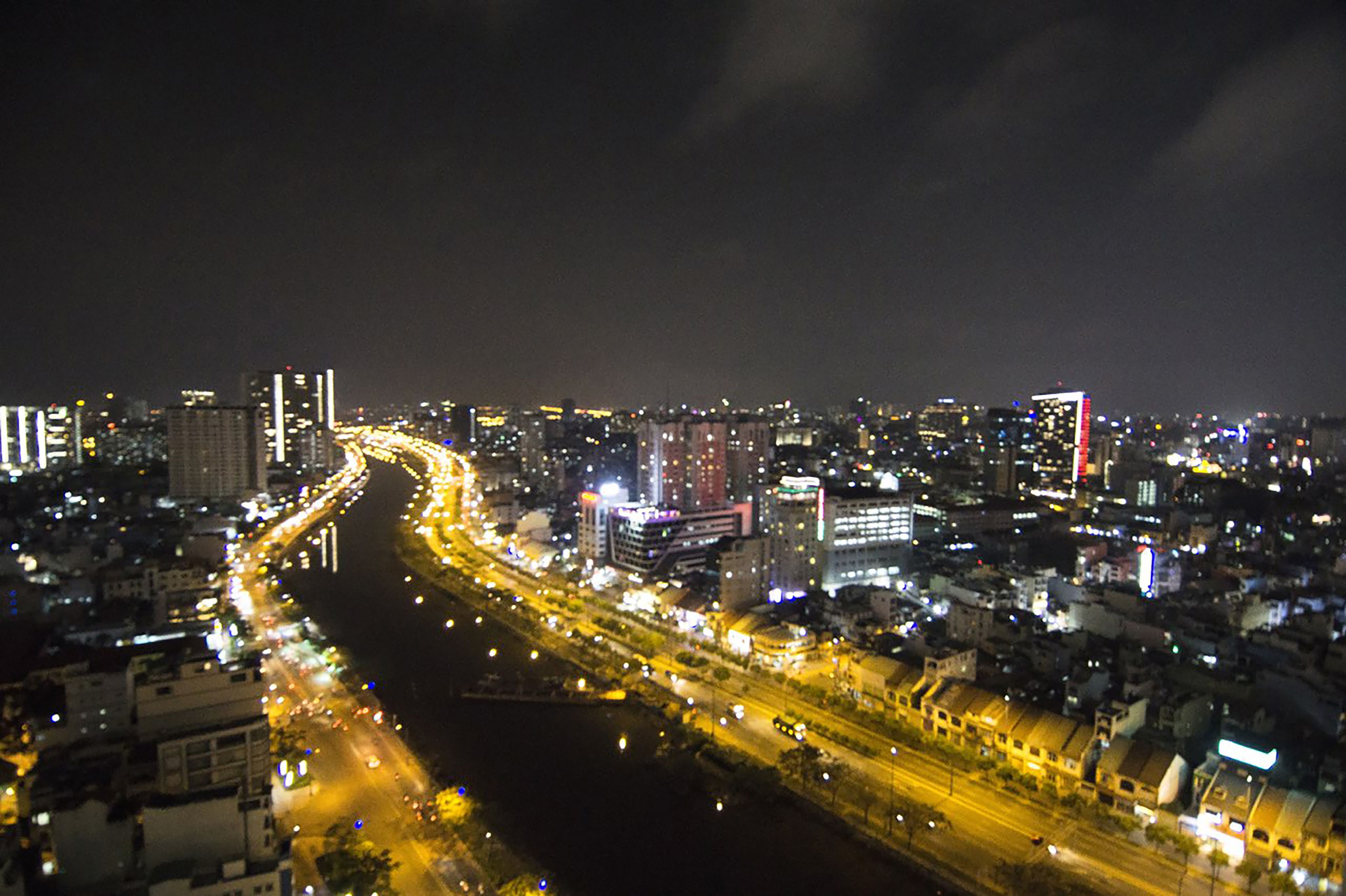 Un paisaje urbano nocturno con un río que lo atraviesa, edificios iluminados y una carretera concurrida con vehículos en movimiento.
