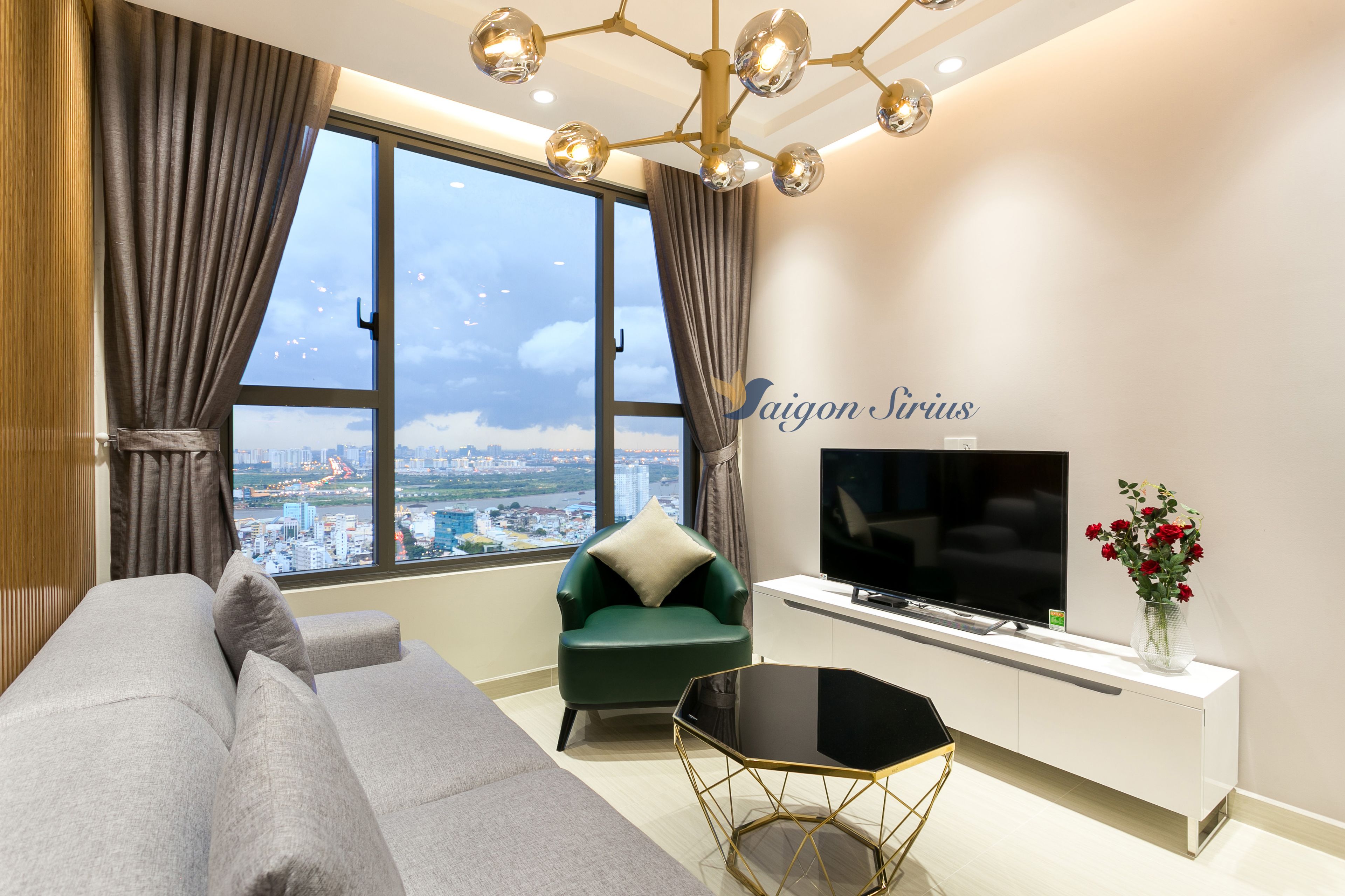 Un salón moderno con un sofá, un sillón verde, un televisor y vistas a la ciudad a través de amplias ventanas.