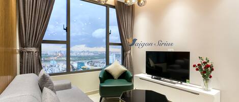 Un salón moderno con un sofá, un sillón verde, un televisor y vistas a la ciudad a través de amplias ventanas.