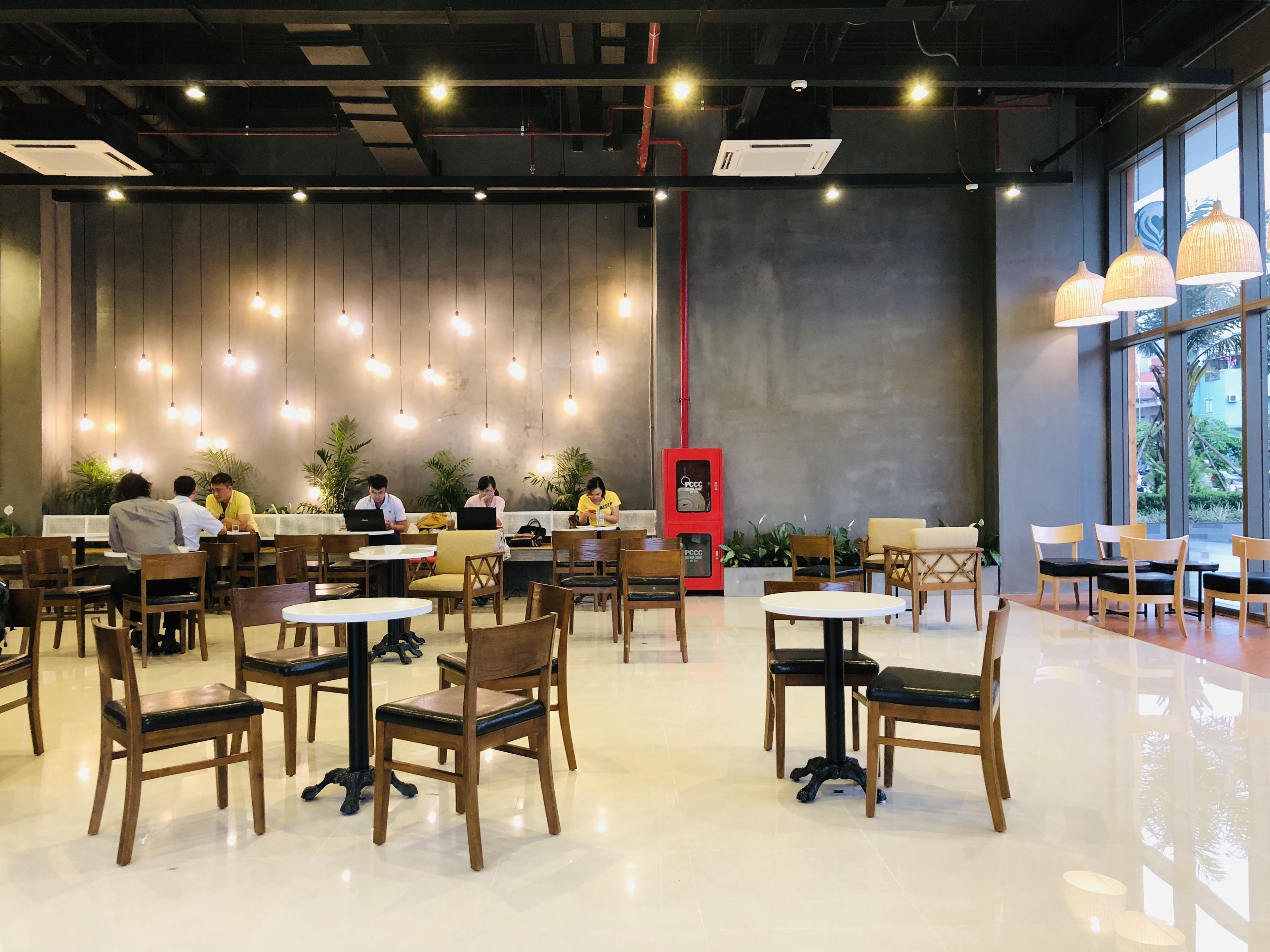 A modern cafe with wooden chairs and tables, a red vending machine, and hanging pendant lights.