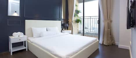 A modern hotel room with a large bed, a bedside table with a lamp, a small table with a clock, and a view of the city through large windows.